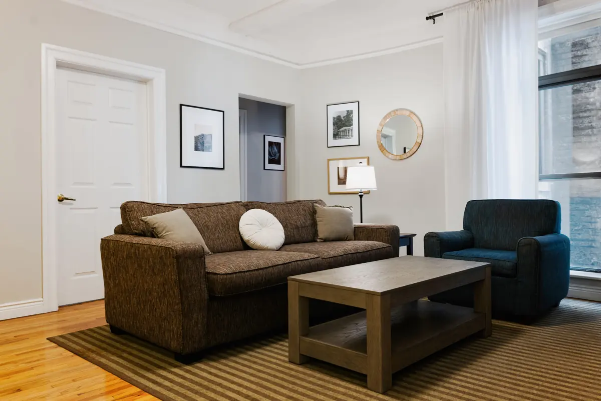 Living room with a brown sofa, blue armchair, wooden coffee table, lamp and framed artwork on light walls.