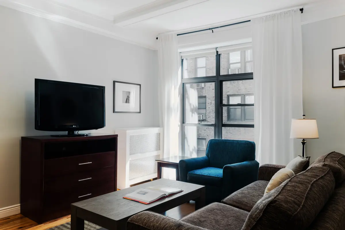 Living room with a TV on a dark cabinet, a blue armchair, a brown sofa, a wooden coffee table and tall windows with white curtains.