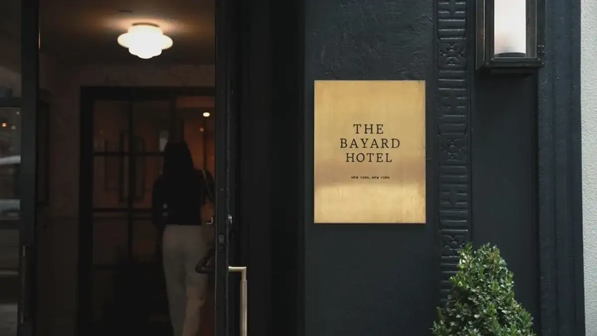 Dark building entrance with a brass plaque reading The Bayard Hotel, New York, and a person walking inside.