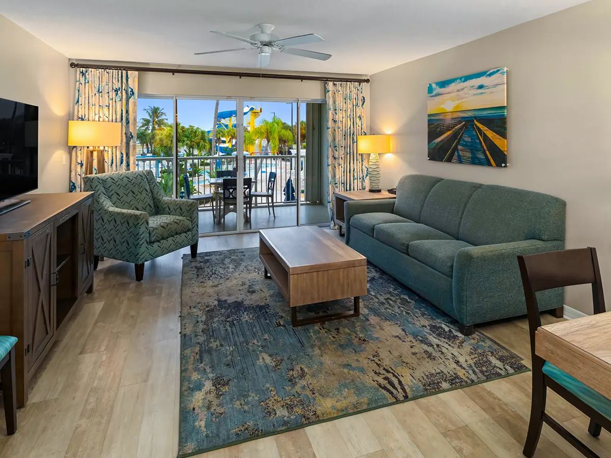 Similar room (illustrative): living room with a teal sofa, patterned armchair, wooden coffee table, TV and a balcony with palm trees outside.