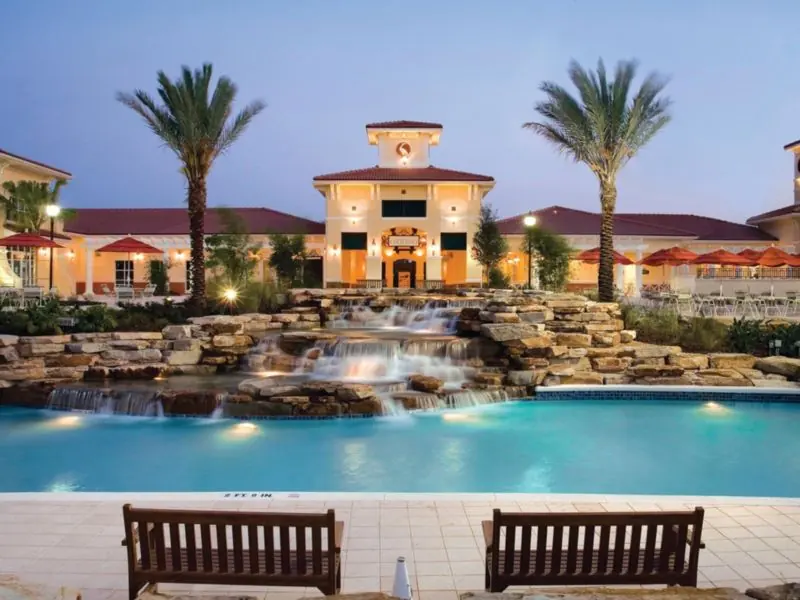 Pool with a rock waterfall and palm trees in front of a resort building, with two benches in the foreground.