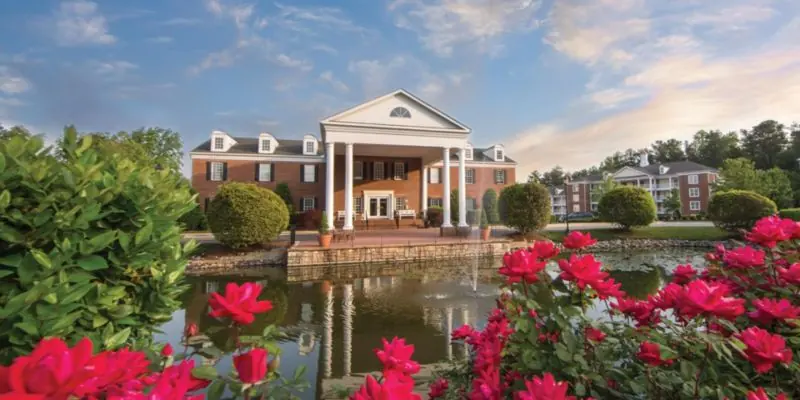 Colonial-style brick building with white columns behind a pond and red flowering bushes.