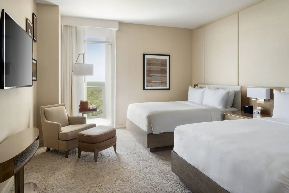 Bedroom with two beds, an armchair with an ottoman, a wall-mounted television and a window.