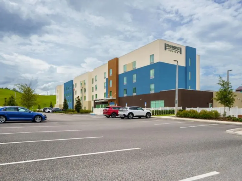 Hotel building with blue and tan sections seen across a parking lot with parked cars.