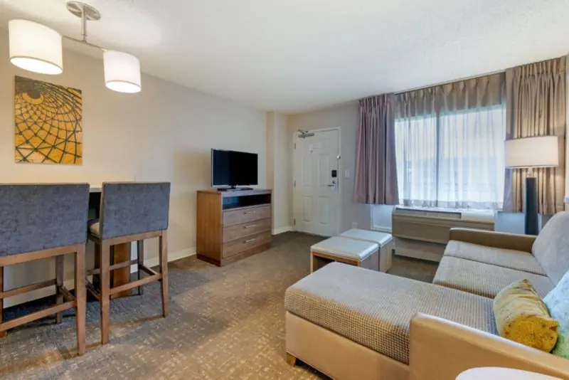Suite living area with a sectional sofa, two bar stools at a kitchen counter, a television on a dresser and a window with curtains.